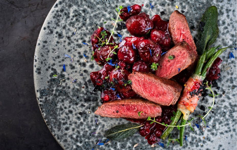 Een grijs gespikkeld bord met daarop stukjes edelhertbiefstuk staat op een donker grijze, houtskool achtige ondergrond. Naast de edelhertbiefstuk ligt een bundeltje sperziebonen die omwikkeld zijn met spek en een saus van rode bessen.
