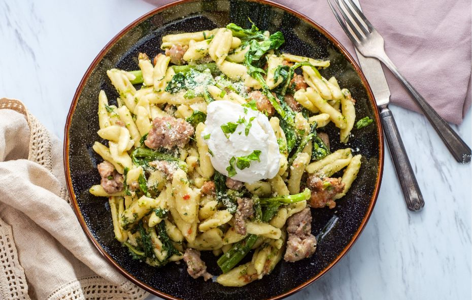 Een donker bord gevuld met pasta, lamsmergeuz stukjes, groene asperges en een bol mozzarella staat op een lichte ondergrond met een katoenen servet. Ernaast ligt een mes en vork.