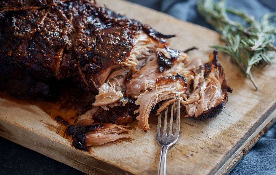 Pulled pork uit de slowcooker op een houten plank met een vork erbij die het vlees uit elkaar trekt.