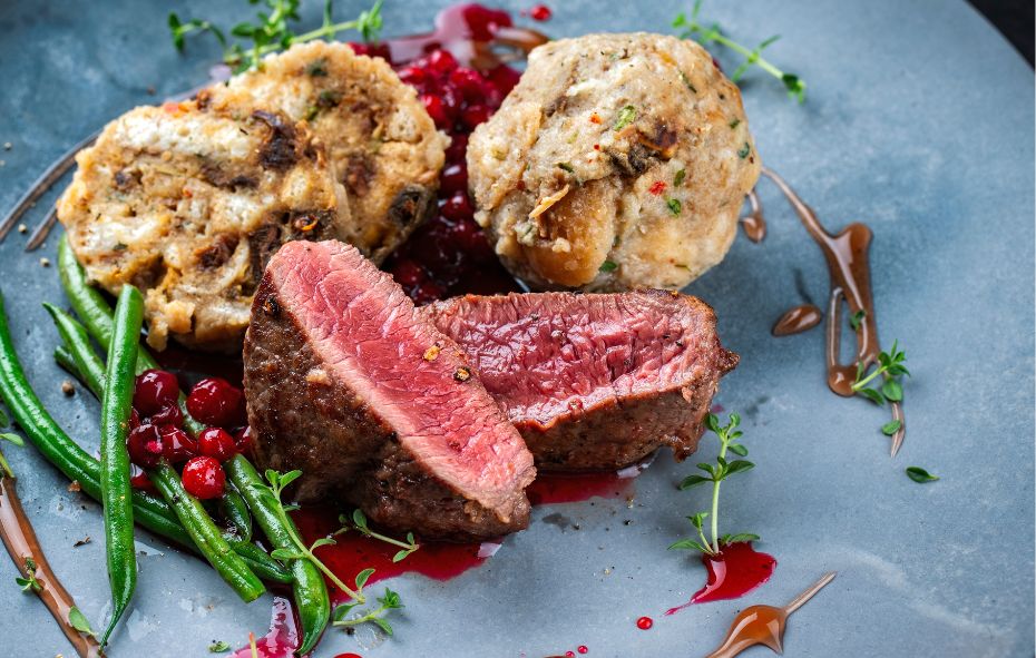 Op een blauwgrijs bord ligt een wild zwijn biefstuk die is doorgesneden. De snijkant ligt naarboven. De biefstuk ligt op een rode saus, ernaast liggen rode bessen, sperzieboontjes en knoedels