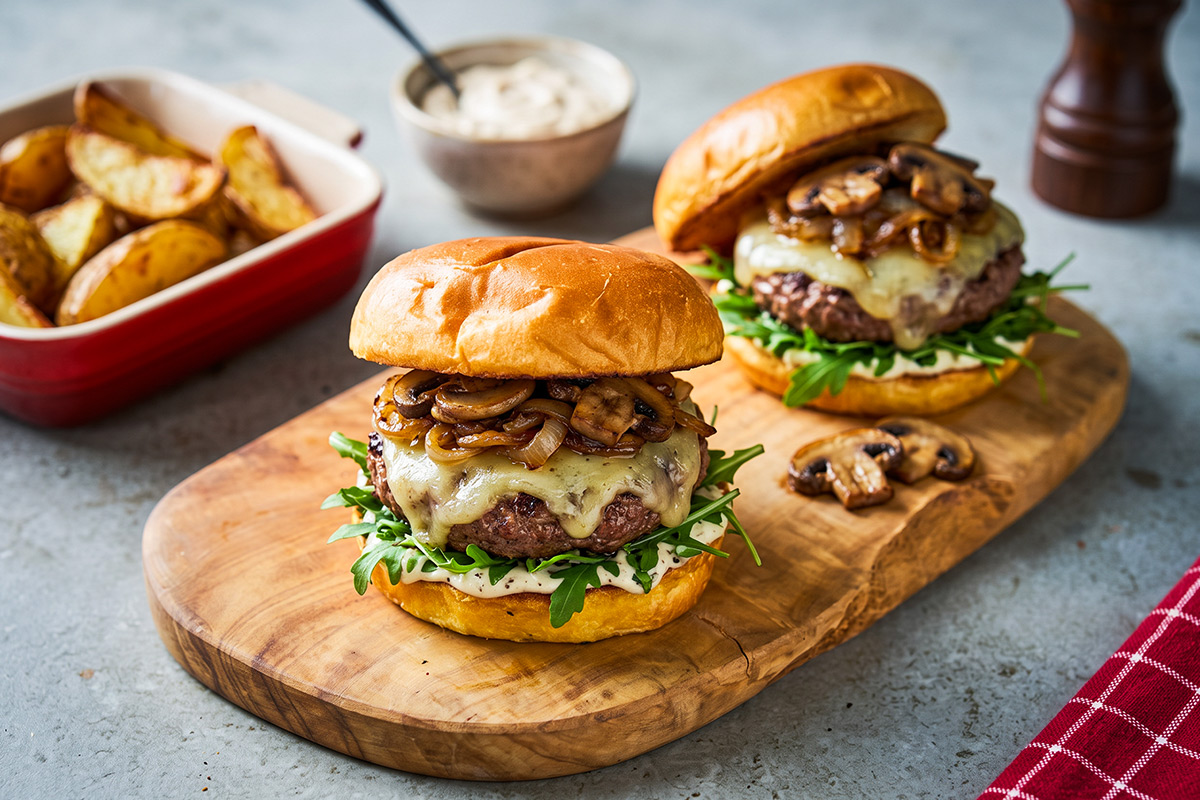 Herfstburger met gesmolten oude kaas, gebakken champignons, gekaramelliseerde rode ui, rucola en truffelmayonaise, met aardappelpartjes op een houten plank