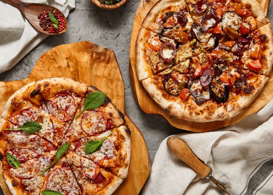 twee versgebakken en zelfgemaakte pizza's op houten planken met daarom heen een lepe met kruiden, theedoek en een pizzasnijder.